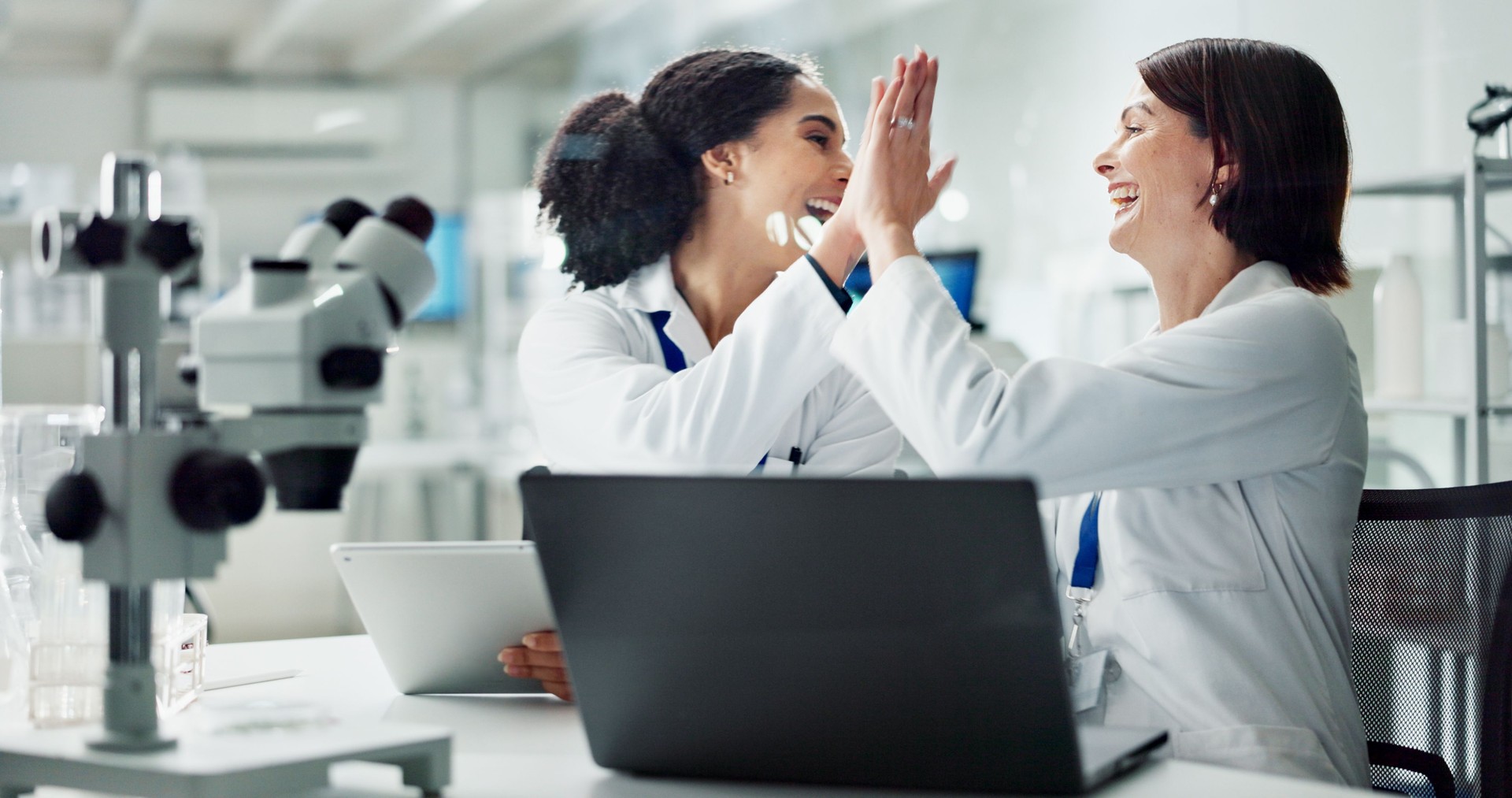 Happy woman, scientist and high five with colleague for teamwork, breakthrough or new discovery on laptop at lab. Female person or medical professionals touching with smile for success at laboratory